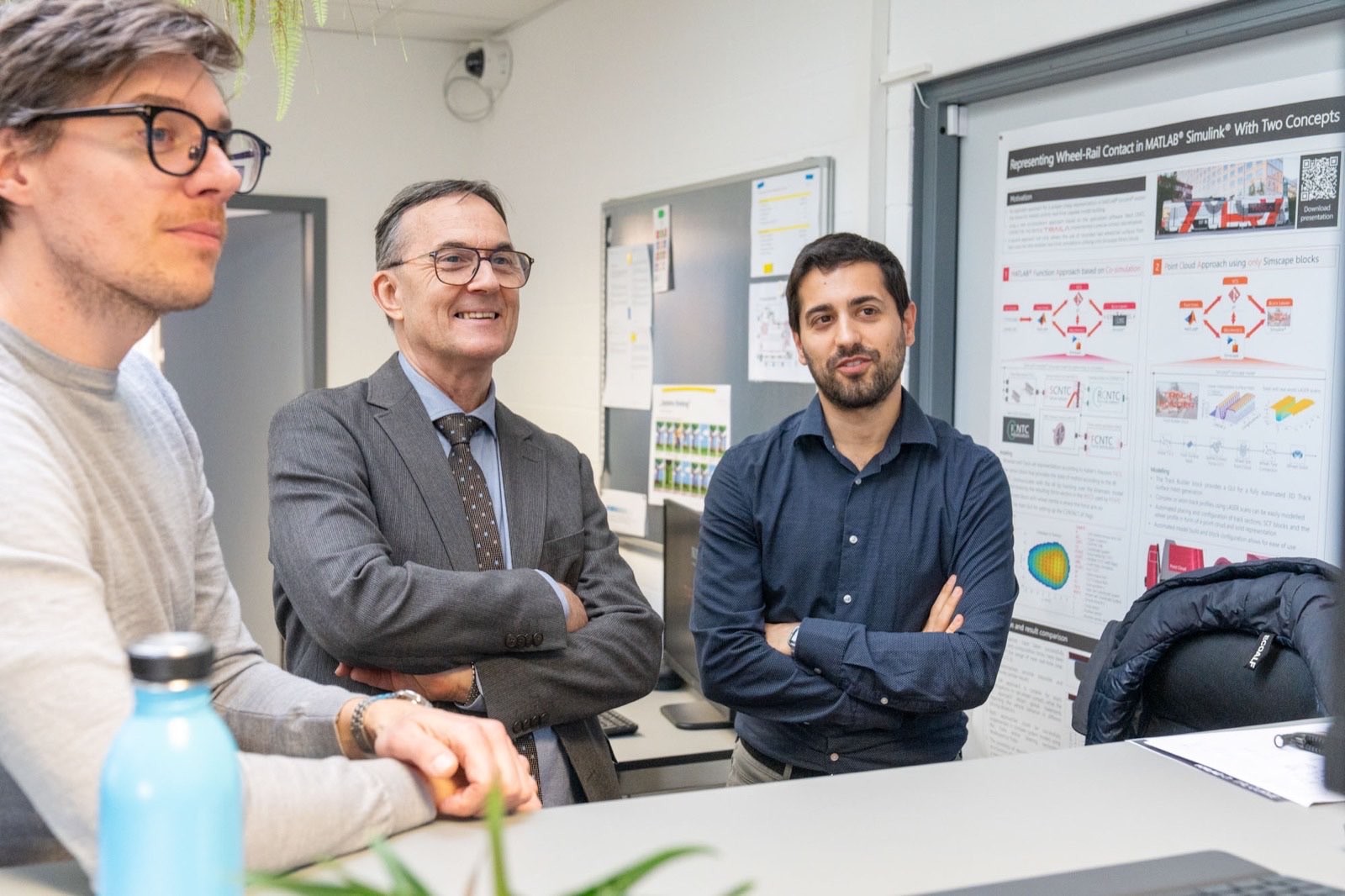 Three colleagues stand by a wheel-rail contact research poster in a Traila office.